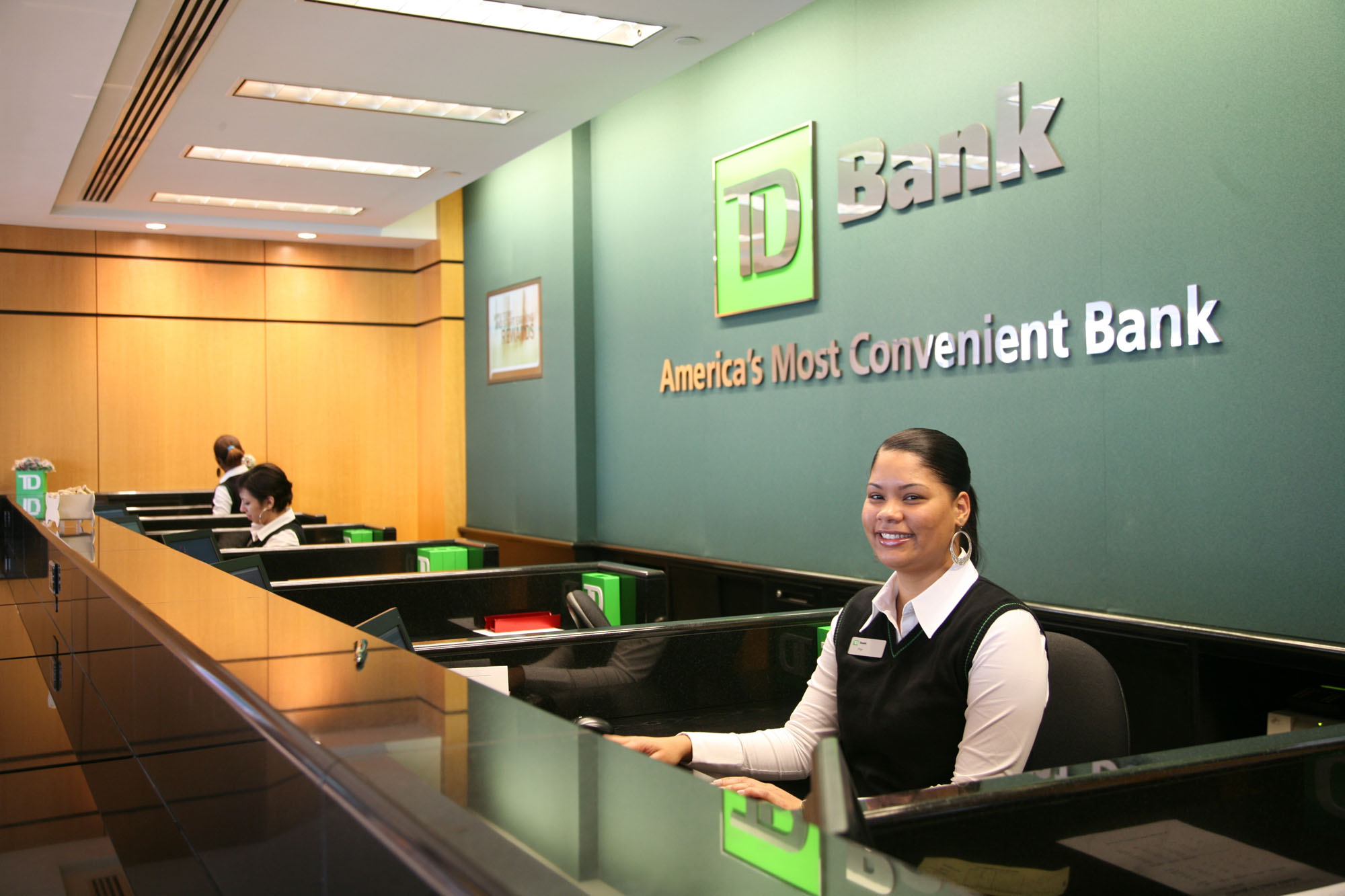  A female bank teller in a green vest and white blouse sits at a counter in a TD Bank branch.